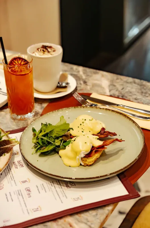 uova alla benedict su un piatto, con accanto un cappuccino ed un succo di frutta